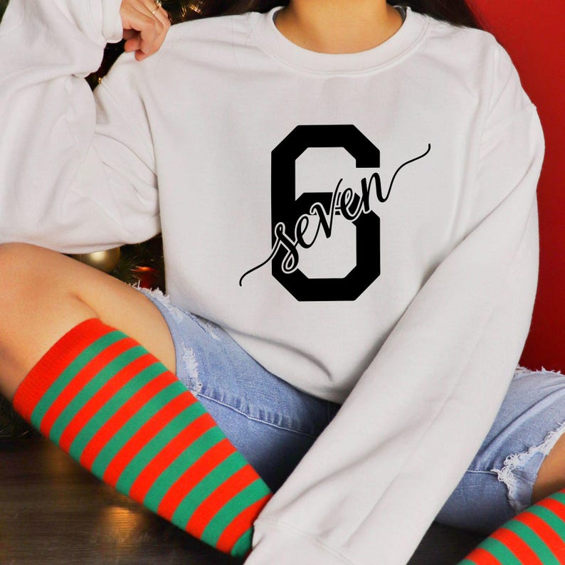May include: White sweatshirt with a large black number 6 and the word "seven" in a cursive font. The person is wearing red and green striped knee-high socks and blue jeans.