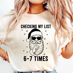 May include: Cream-colored t-shirt with a graphic of Santa Claus wearing sunglasses and a black hat, with his finger over his lips. The text reads "CHECKING MY LIST 6-7 TIMES" in black font, with snowflake and dot accents.
