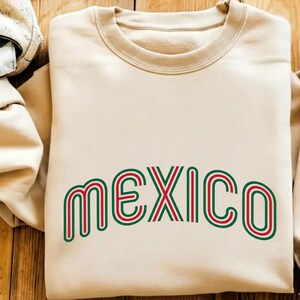 May include: A beige crewneck sweatshirt featuring the word MEXICO printed in a stylized, rounded font. The lettering consists of concentric lines in alternating shades of red and green, creating a layered, striped effect across the chest.