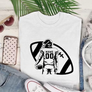 以下が含まれることがあります： 白いTシャツ。黒いグラフィックで、アメフト選手がボールを持っています。ジャージには数字「00」と「NAME」の文字。デザインは黒いアメフトの形に配置されています。