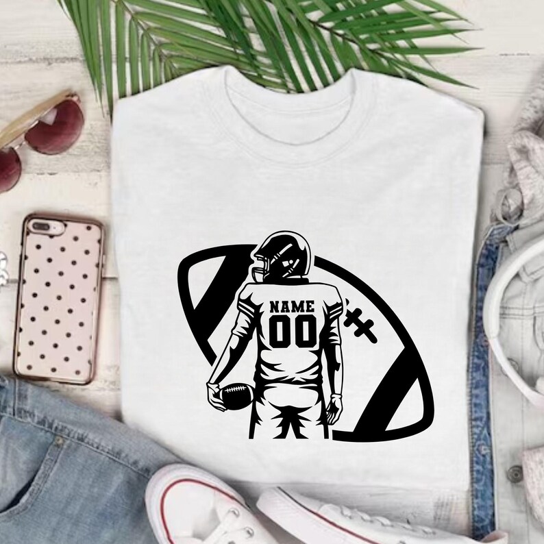 以下が含まれることがあります： 白いTシャツ。黒いグラフィックで、アメフト選手がボールを持っています。ジャージには数字「00」と「NAME」の文字。デザインは黒いアメフトの形に配置されています。