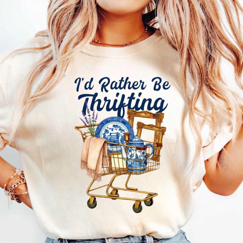 Może przedstawiać: Kremowa koszulka z niebieskim napisem "I'd Rather Be Thrifting". Grafika w&oacute;zka sklepowego wypełniona jest niebiesko-białą porcelaną, oprawionym zdjęciem i szalikiem. W&oacute;zek jest ze złotego metalu.
