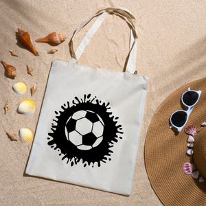 Pu&ograve; includere: Una borsa tote beige con un design a pallone da calcio in bianco e nero. Il pallone da calcio &egrave; circondato da una grafica a spruzzo nera. La borsa ha due manici beige. L'immagine include anche un cappello di paglia e occhiali da sole.
