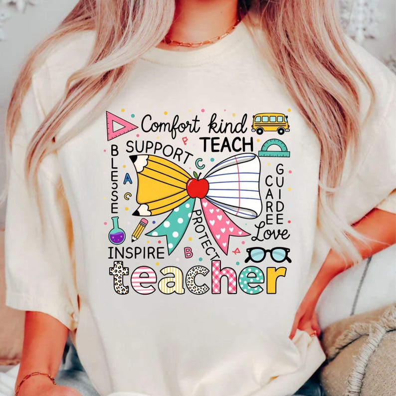 May include: Cream-colored t-shirt with a colorful graphic design for teachers. The design includes a pencil, apple, and bow, with words like "Comfort," "Teach," and "Teacher." A school bus and glasses are also included.