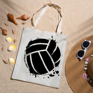Op de afbeelding: Een beige draagtas met een groot zwart volleybalontwerp. De tas heeft witte handvatten en ligt op zand, naast een strohoed en schelpen. Het volleybalontwerp heeft een distressed, splatter effect.
