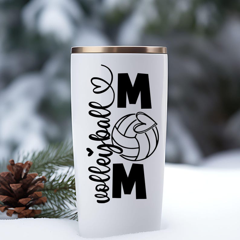 May include: White insulated tumbler with a gold rim. The tumbler features the words "volleyball mom" in black script and block letters, along with a volleyball graphic with a heart. The background is a snowy outdoor scene.