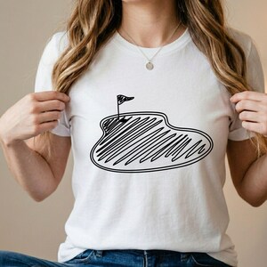 May include: White t-shirt featuring a black line drawing of a golf green with a flag. The design is a simple, hand-drawn style. The shirt is made of a soft material and is suitable for casual wear. The shirt is a great gift for golf enthusiasts.