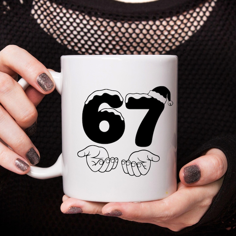 May include: White ceramic mug featuring the number 67 in black, with a Santa hat on the 7. The mug also has two hands with open palms below the numbers. The mug is held by a person with dark nail polish.
