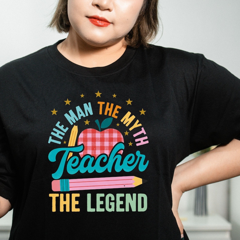 May include: Black t-shirt with a colorful graphic design. The design includes an apple with a red and white checkered pattern, pencils, stars, and the text "THE MAN THE MYTH Teacher THE LEGEND".