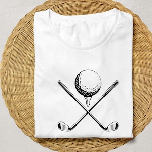 May include: White t-shirt featuring a black graphic of a golf ball on a tee with two crossed golf clubs. The shirt is folded on a woven, light brown surface. The design is simple and bold, perfect for golf enthusiasts.