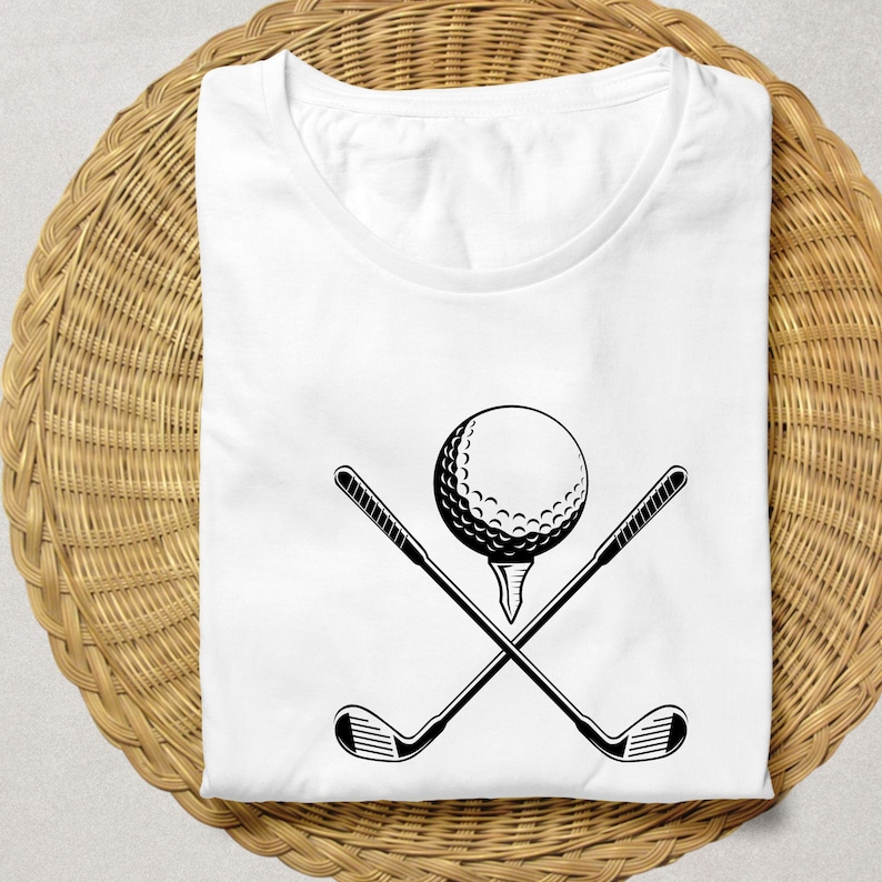 May include: White t-shirt featuring a black graphic of a golf ball on a tee with two crossed golf clubs. The shirt is folded on a woven, light brown surface. The design is simple and bold, perfect for golf enthusiasts.