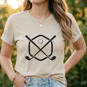 May include: Beige t-shirt featuring a black golf-themed graphic. The graphic includes crossed golf clubs, a golf ball, and a shield-shaped outline. The shirt has short sleeves and a crew neck.