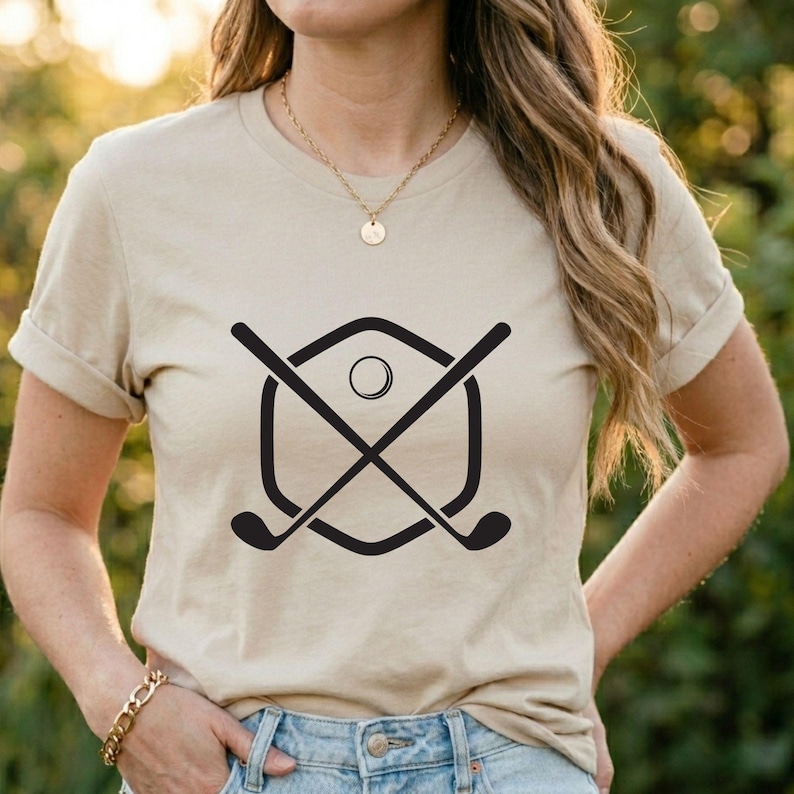 May include: Beige t-shirt featuring a black golf-themed graphic. The graphic includes crossed golf clubs, a golf ball, and a shield-shaped outline. The shirt has short sleeves and a crew neck.