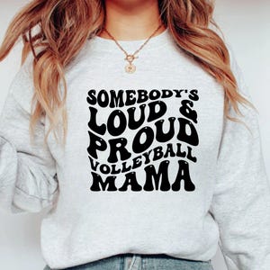 May include: A light gray sweatshirt with the text "SOMEBODY'S LOUD & PROUD VOLLEYBALL MAMA" in a black, bold, retro font. The sweatshirt has long sleeves and a crew neck.