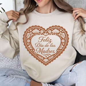 May include: A beige sweatshirt featuring a large, heart-shaped design in shades of brown. The heart is filled with floral and decorative elements and the words "Feliz D&iacute;a de las Madres" are written in a script font.