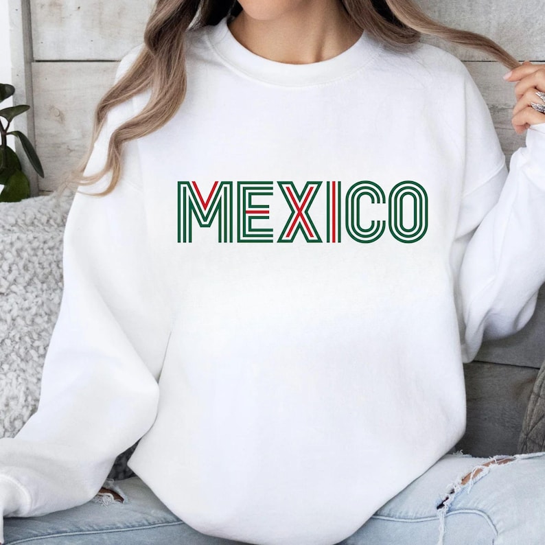 May include: A white long-sleeve crewneck sweatshirt featuring the word MEXICO printed in a stylized font. The letters are composed of multiple parallel lines in dark green, with the letter X accented by a single red line through the center.