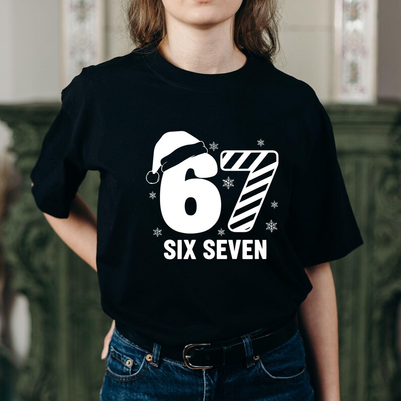 May include: Black t-shirt with a white graphic featuring the numbers "67" with a Santa hat on the "6" and a candy cane design for the "7". The words "SIX SEVEN" are printed below the numbers, with snowflakes around the numbers.