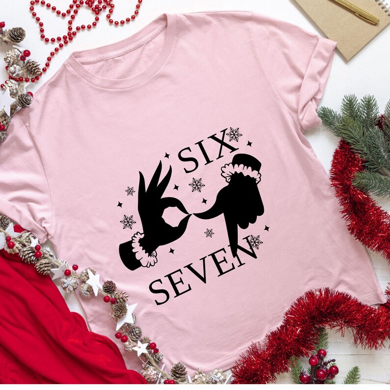 May include: A light pink t-shirt with a black graphic design. The design features two hands forming the number six and seven with the text "SIX" and "SEVEN" in a decorative font. Snowflakes and stars surround the hands.