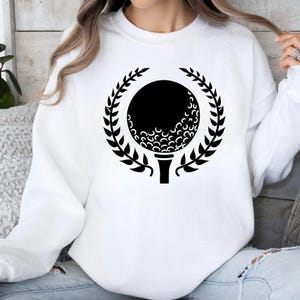 May include: White sweatshirt featuring a black golf ball design within a laurel wreath. The golf ball is on a tee, and the design is centered on the front of the shirt. A casual, sporty apparel item.