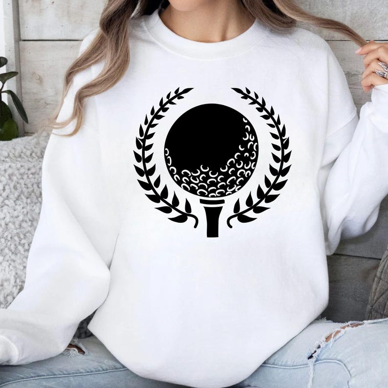 May include: White sweatshirt featuring a black golf ball design within a laurel wreath. The golf ball is on a tee, and the design is centered on the front of the shirt. A casual, sporty apparel item.