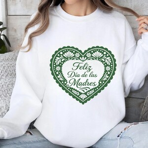 May include: White sweatshirt featuring a green heart-shaped design with floral patterns and the text "Feliz D&iacute;a de las Madres". The design is centered on the front of the sweatshirt.