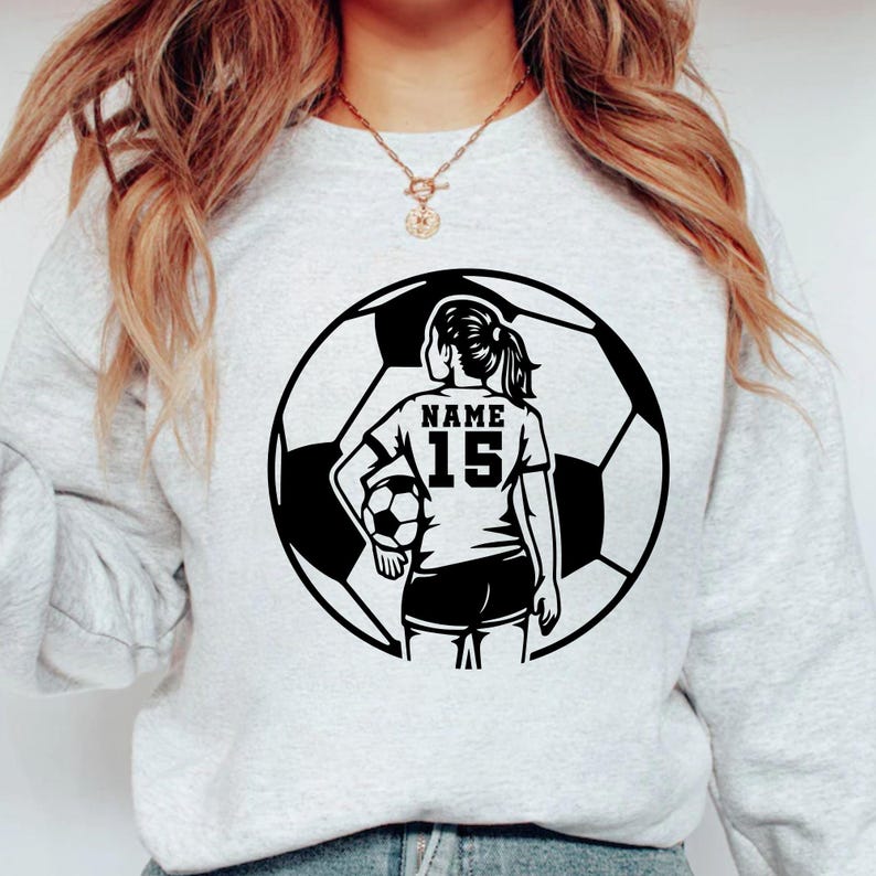 Pu&ograve; includere: Una felpa grigio chiaro con una grafica in bianco e nero di una calciatrice che tiene un pallone da calcio. La giocatrice &egrave; all'interno di un contorno di pallone da calcio. La maglia della giocatrice ha il testo "NAME" e il numero "15".
