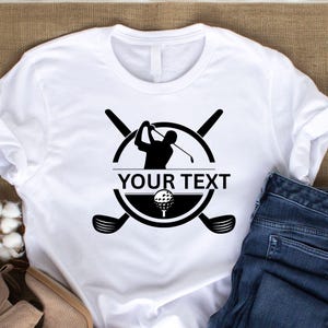 May include: White t-shirt featuring a black golf-themed design. The design includes a golfer silhouette, golf ball, and crossed golf clubs, with the text "YOUR TEXT" in a bold font. The shirt is paired with blue jeans.