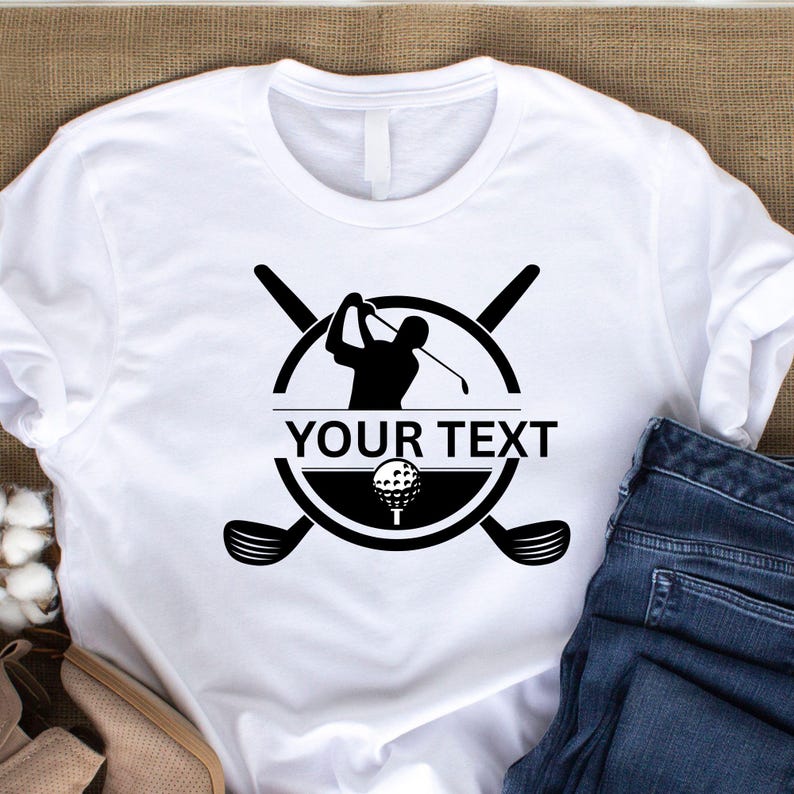 May include: White t-shirt featuring a black golf-themed design. The design includes a golfer silhouette, golf ball, and crossed golf clubs, with the text "YOUR TEXT" in a bold font. The shirt is paired with blue jeans.