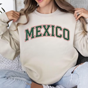 May include: A beige crewneck sweatshirt featuring the word MEXICO printed in an arched collegiate font. The lettering is dark green with a thin red outline. The garment has long sleeves and a ribbed collar, cuffs, and hemline.