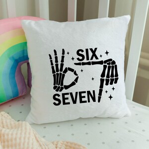 May include: White plush pillow with a black skeleton hand design. The design spells out "SIX SEVEN" with the fingers forming the numbers. Small star shapes are scattered around the text.