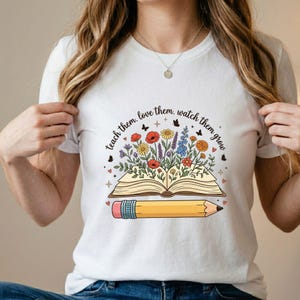 May include: White t-shirt featuring a colorful illustration of an open book with flowers and butterflies, and a yellow pencil. The text "teach them. love them. watch them grow" is arched above the book.