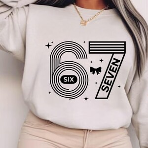 May include: Cream-colored sweatshirt featuring a black graphic design of the numbers "67". The number "6" is stylized with concentric circles and the word "SIX" inside. The number "7" is in a bold, angled font with the word "SEVEN".