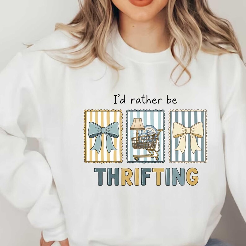 Może przedstawiać: Biała bluza z napisem "I'd rather be thrifting". Projekt zawiera trzy oprawione obrazy kokard i w&oacute;zka sklepowego wypełnionego przedmiotami. Słowo "THRIFTING" jest wydrukowane dużymi literami pod obrazami.