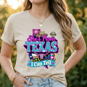 May include: A beige t-shirt with a graphic design featuring the text "She's From TEXAS I Can Tell" in a turquoise and purple color scheme. The design includes a map of Texas, a cowboy hat, and other Texas-themed elements.