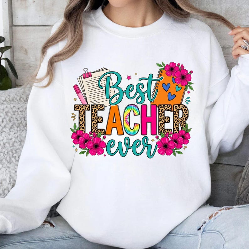 May include: White sweatshirt with a colorful graphic that reads "Best Teacher Ever." The design includes a leopard print, flowers, and a tie-dye effect. A notepad and pen are also featured in the design.
