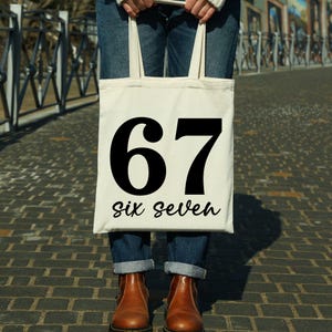 May include: A cream-colored tote bag with the number "67" in bold black font and the words "six seven" in a script font. The bag is held by a person wearing blue jeans and brown boots.