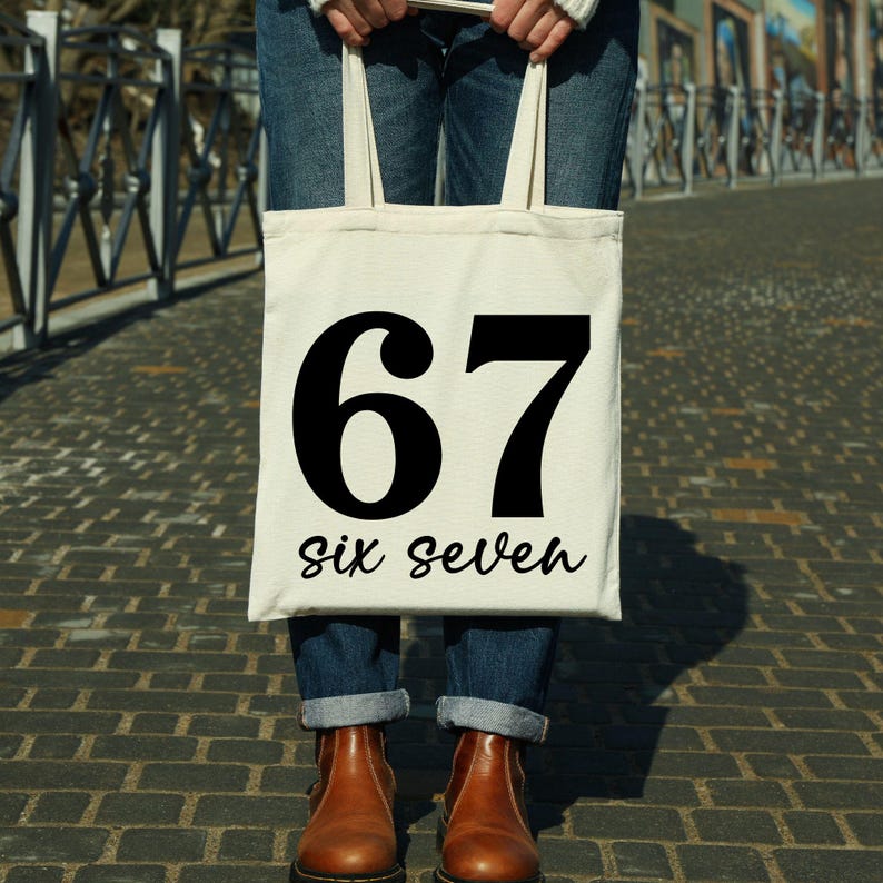 May include: A cream-colored tote bag with the number "67" in bold black font and the words "six seven" in a script font. The bag is held by a person wearing blue jeans and brown boots.