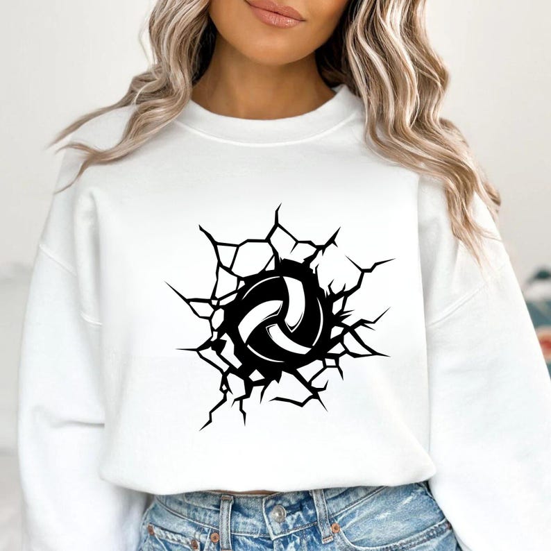 May include: White sweatshirt featuring a black and white volleyball design. The volleyball graphic appears to be breaking through a cracked surface, creating a dynamic visual effect. The sweatshirt is a casual, sporty apparel item.