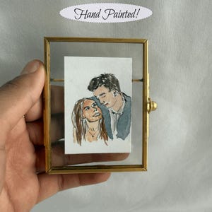 May include: A hand-painted watercolor portrait of a couple in a gold-toned frame. The artwork features a man and woman gazing at each other. The frame is rectangular with a glass front. Text above the frame reads "Hand Painted!"