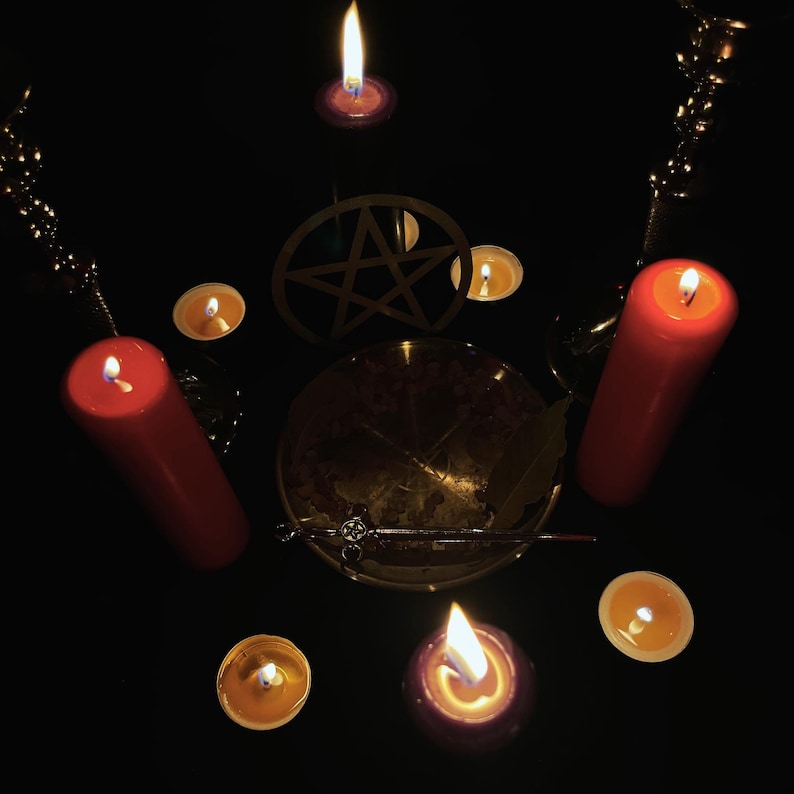 May include: A ritualistic arrangement featuring lit candles of varying sizes and colors, including red pillar candles and small tealights. A pentagram and a bowl are at the center, with a wand and other items, all set against a dark background.