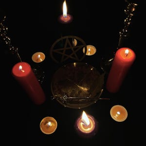 K&ouml;nnte beinhalten: Eine dunkle, rituelle Anordnung mit brennenden Kerzen in verschiedenen Gr&ouml;&szlig;en und Farben. Rote S&auml;ulenkerzen und kleine Teelichter umgeben eine zentrale Schale mit Kr&auml;utern, einem Pentagramm und einem Zauberstab. Dekorative Ketten.