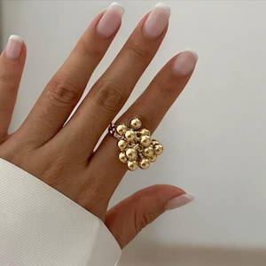 Puede incluir: Un anillo dorado con un racimo de pequeñas cuentas redondas. El anillo se lleva en un dedo, con la mano y parte de una manga blanca visibles. El diseño del anillo se asemeja a un racimo de uvas.