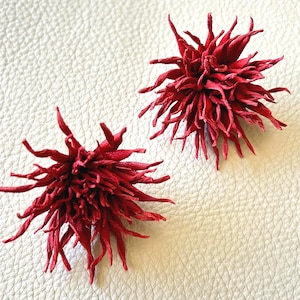 May include: Two handmade, bright red, spiky flower-shaped ornaments. The ornaments have a textured, layered appearance, with numerous thin, pointed petals radiating outwards. They are set against a white, textured background.