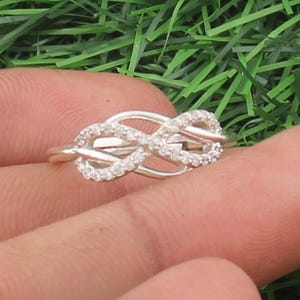 May include: A silver ring featuring a knot design, adorned with small, clear gemstones. The ring is displayed against a backdrop of green grass, highlighting its intricate details and sparkling stones.