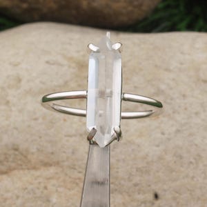 Op de afbeelding: Een zilveren ring met een heldere, rechthoekige kristal. Het kristal is verticaal geplaatst in een minimalistisch ontwerp. De ring wordt vastgehouden door een zilveren gereedschap, dat de details van de ring tegen een neutrale achtergrond benadrukt.