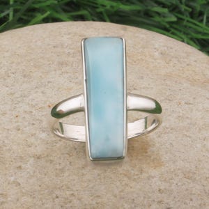 May include: A rectangular, light blue gemstone ring with a silver band. The gemstone is set vertically in a silver bezel. The ring is displayed on a light brown stone, with green grass in the background.