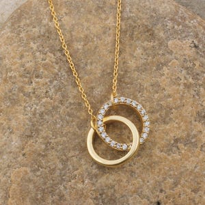 Pode incluir: Um colar dourado com dois círculos interligados. Um círculo é adornado com pequenas pedras preciosas transparentes, enquanto o outro é de cor dourada sólida. O colar é exibido em uma pedra texturizada marrom clara, com grama verde ao fundo.