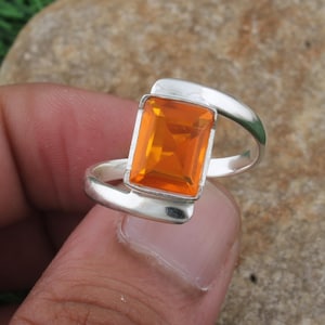 May include: A silver ring featuring a rectangular, orange gemstone. The gemstone is set in a modern, minimalist design with a curved band. The ring is held in a hand, showcasing the bright color of the stone.