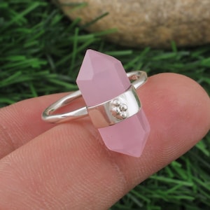 Puede incluir: Anillo de plata con un cristal facetado de color rosa claro. El cristal está sujeto por una banda de plata con un pequeño grupo decorativo de tres círculos. El anillo está sobre un fondo verde.