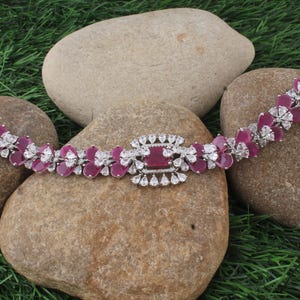 Könnte beinhalten: Ein silberfarbenes Armband mit einem rechteckigen, rubinroten Mittelstein, umgeben von kleineren weißen Steinen. Das Armband hat ein Blumendesign mit rubinroten Blütenblättern und weißen Akzenten. Der Schmuck wird auf glatten, hellbraunen Steinen vor grünem Hintergrund präsentiert.